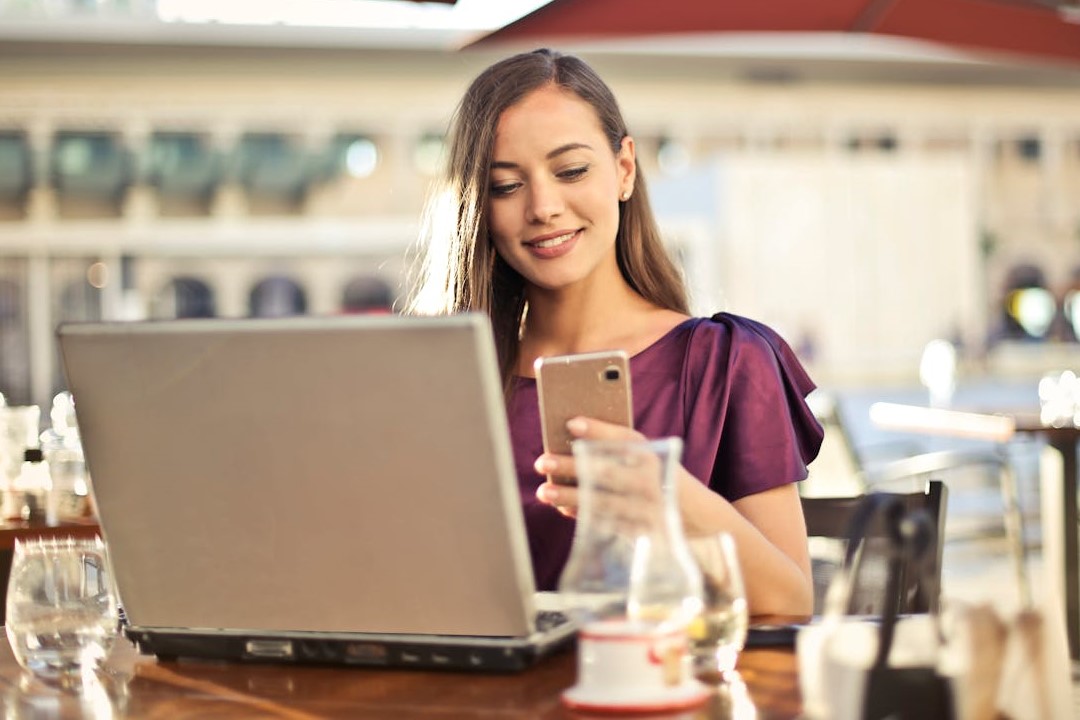 Eine Frau sitzt in einem Café vor ihrem Laptop, während sie auf ihr Smartphone schaut.