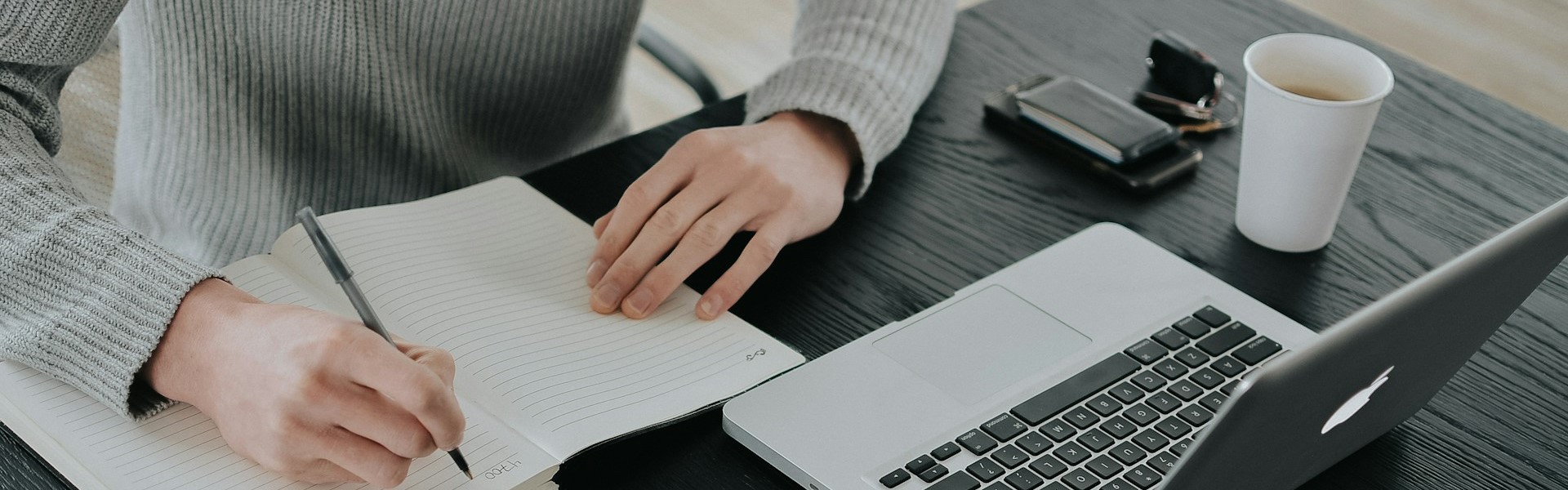 Eine Person sitzt vor dem Laptop mit Stift und Notizbuch.