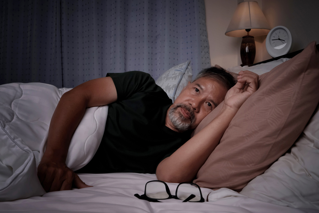 Älterer Mann liegt im Bett und ist gestresst, da er unter Schlaflosigkeit leidet. Es ist dunkel und im Hintergrund ist ein Wecker zu sehen, der 3:45 Uhr anzeigt. 