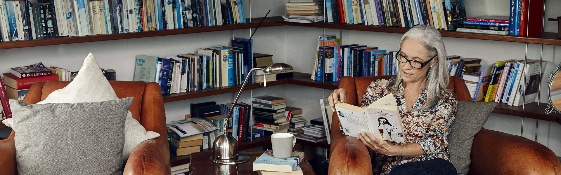 Ältere Frau sitzt im Sessel und liest, hinter ihr mehrere Regale mit Büchern.