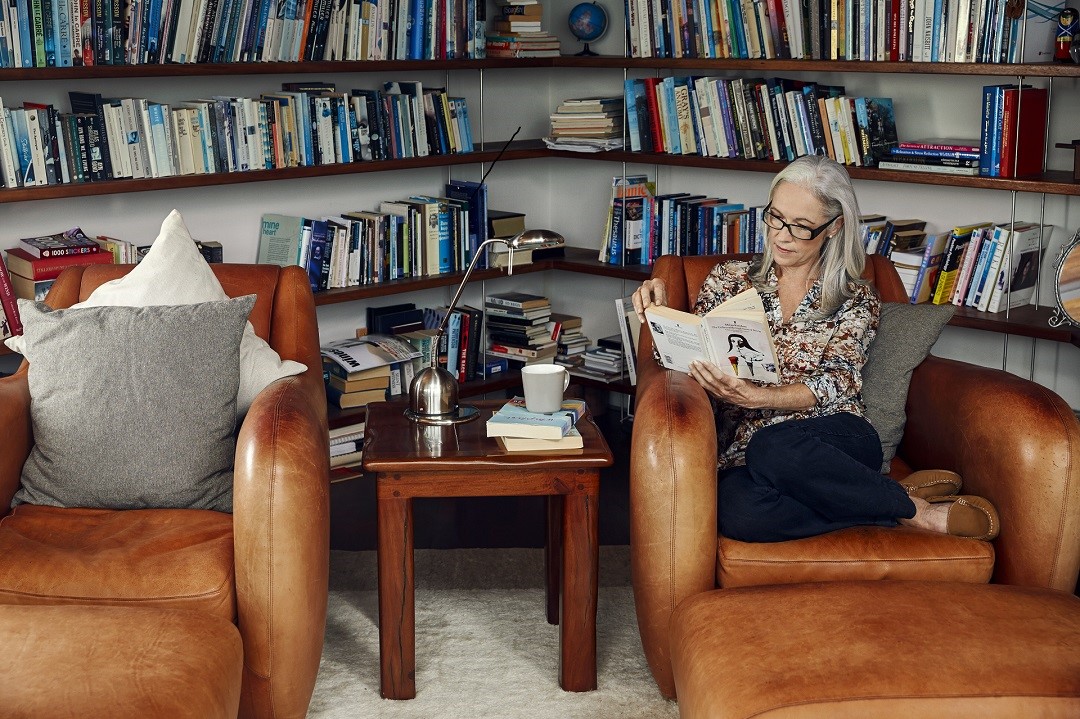 Ältere Frau sitzt im Sessel und liest, hinter ihr mehrere Regale mit Büchern.