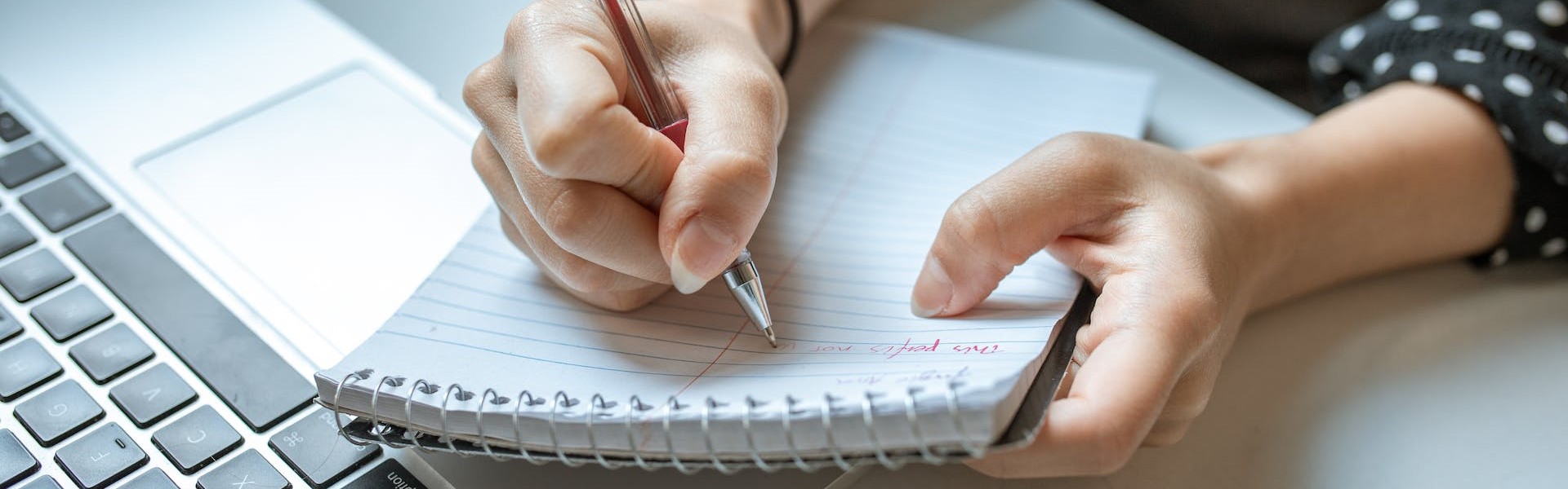 Nahaufnahme einer Person am Laptop, die sich auf einen Block Notizen macht.